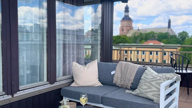 A cozy balcony features a grey couch with pillows, a table holding drinks and grapes, overlooking a cityscape with a historical building in the background under a blue sky.