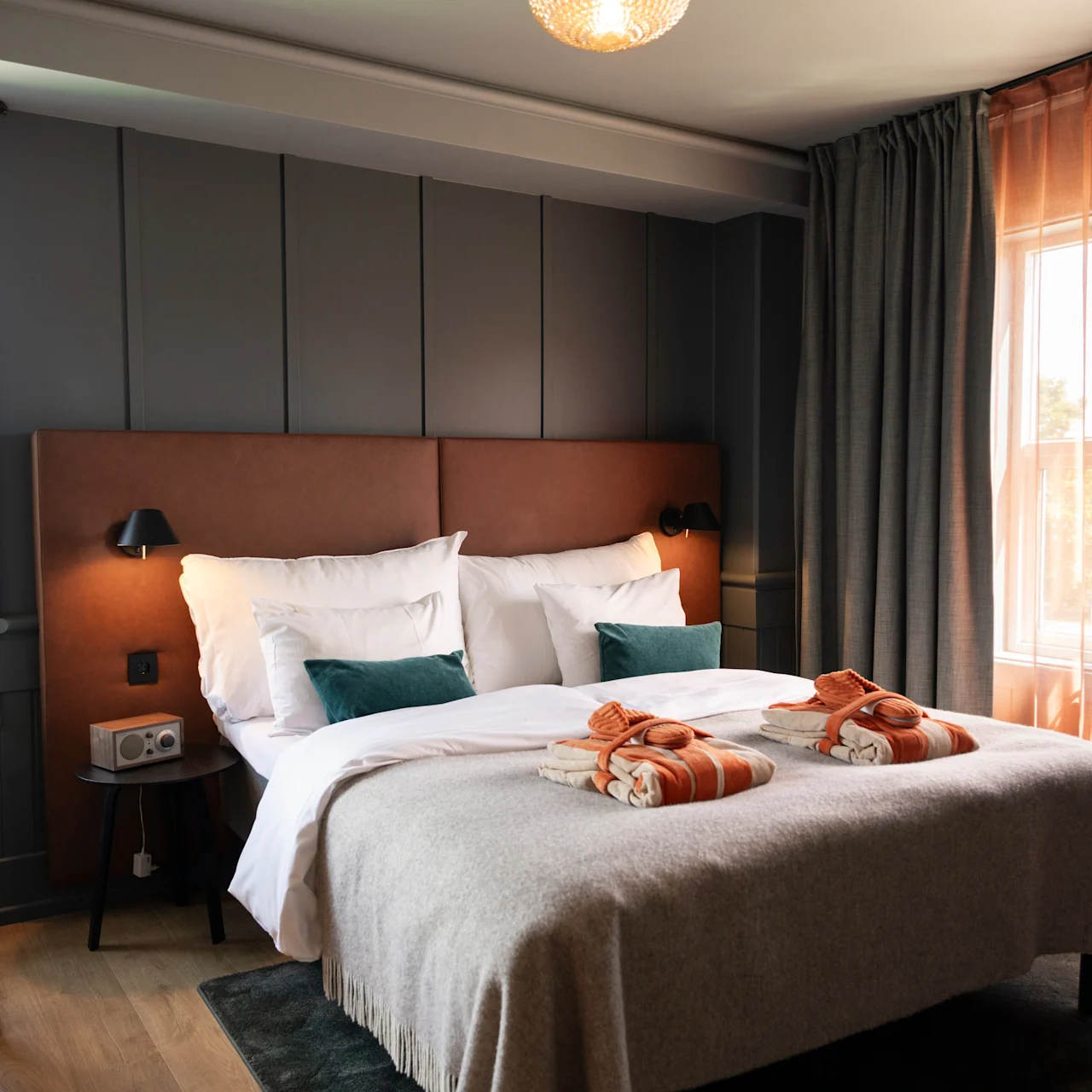 En seng med orange håndklæder og grønne puder står i et moderne hotelværelse med et skrivebord, store vinduer og blødt lys.