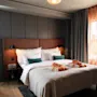 En seng med orange håndklæder og grønne puder står i et moderne hotelværelse med et skrivebord, store vinduer og blødt lys.