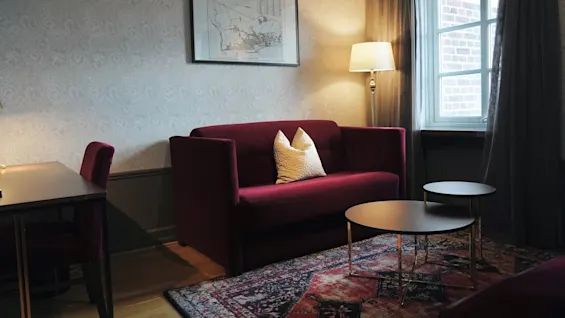 En vinrød sofa med to puter står under innramet kartkunst, ved siden av en tent lampe og et vindu med gardiner; nært er et mønstret teppe og nestede runde bord.