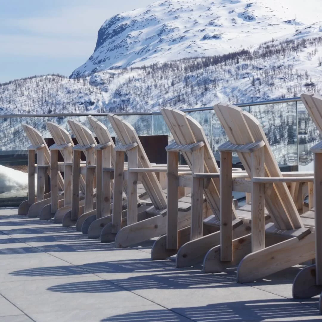 Wooden chairs are aligned on a terrace, casting shadows. They are facing a snow-covered mountain under a clear blue sky.
