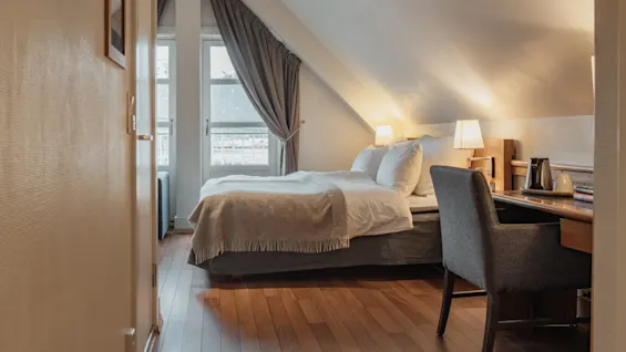 A cozy attic bedroom with a comfortable bed, a window with curtains, and a functional desk area with a chair and warm lighting.