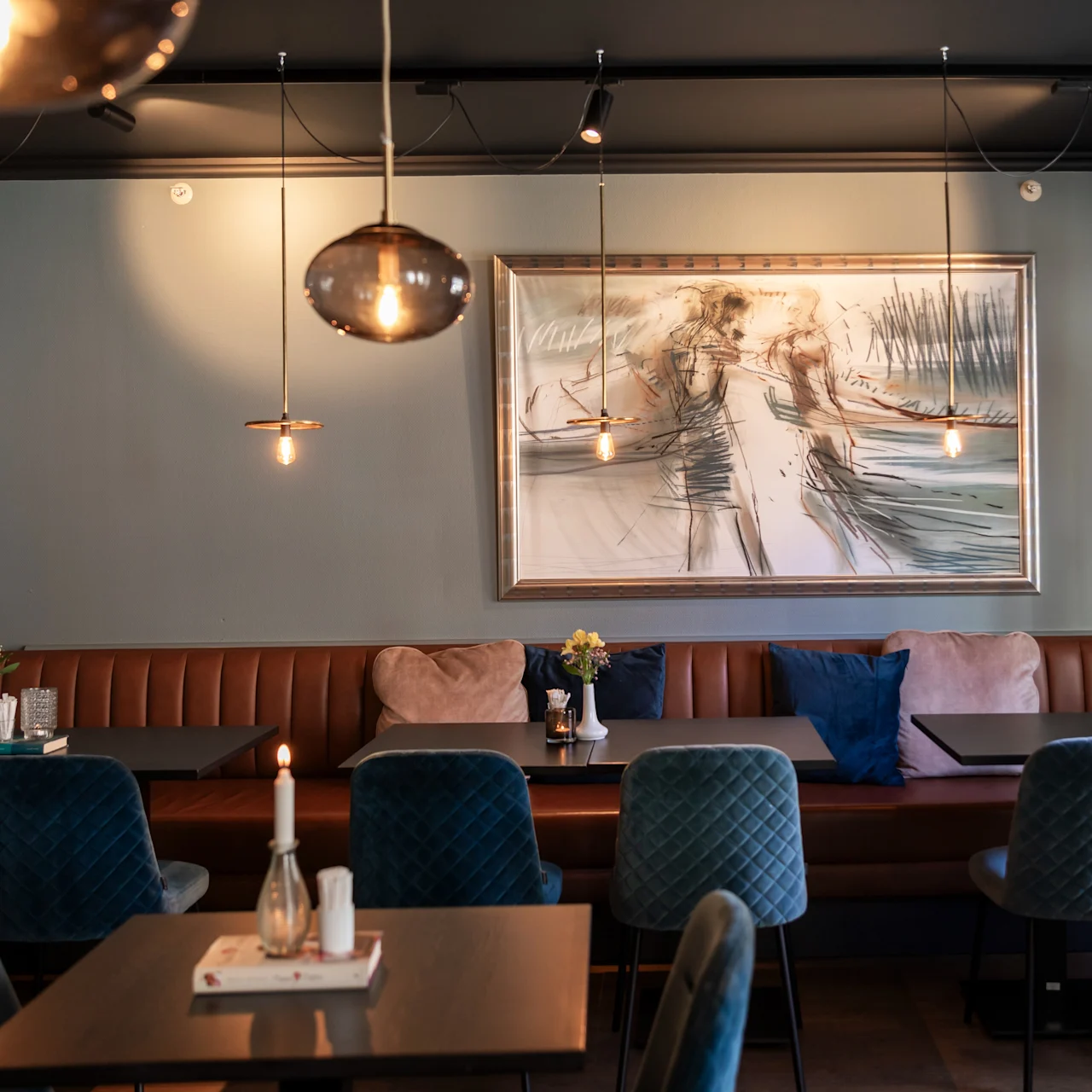 Moderne caféinteriør med en lang brun læderbænk, teal farvede stole, træborde og hængende pendellamper. Et stort abstrakt maleri pryder væggen og skaber en hyggelig atmosfære.