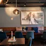 Moderne caféinteriør med en lang brun læderbænk, teal farvede stole, træborde og hængende pendellamper. Et stort abstrakt maleri pryder væggen og skaber en hyggelig atmosfære.