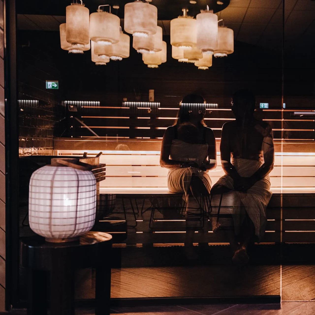 Two people wrapped in towels sit on a wooden sauna bench, surrounded by warm lighting and glass surfaces, with hanging lamps above.