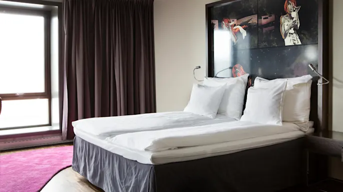 A neatly made double bed with white linens stands in a modern hotel room. The backdrop features dark curtains and a large illustrated headboard with a red-haired figure.
