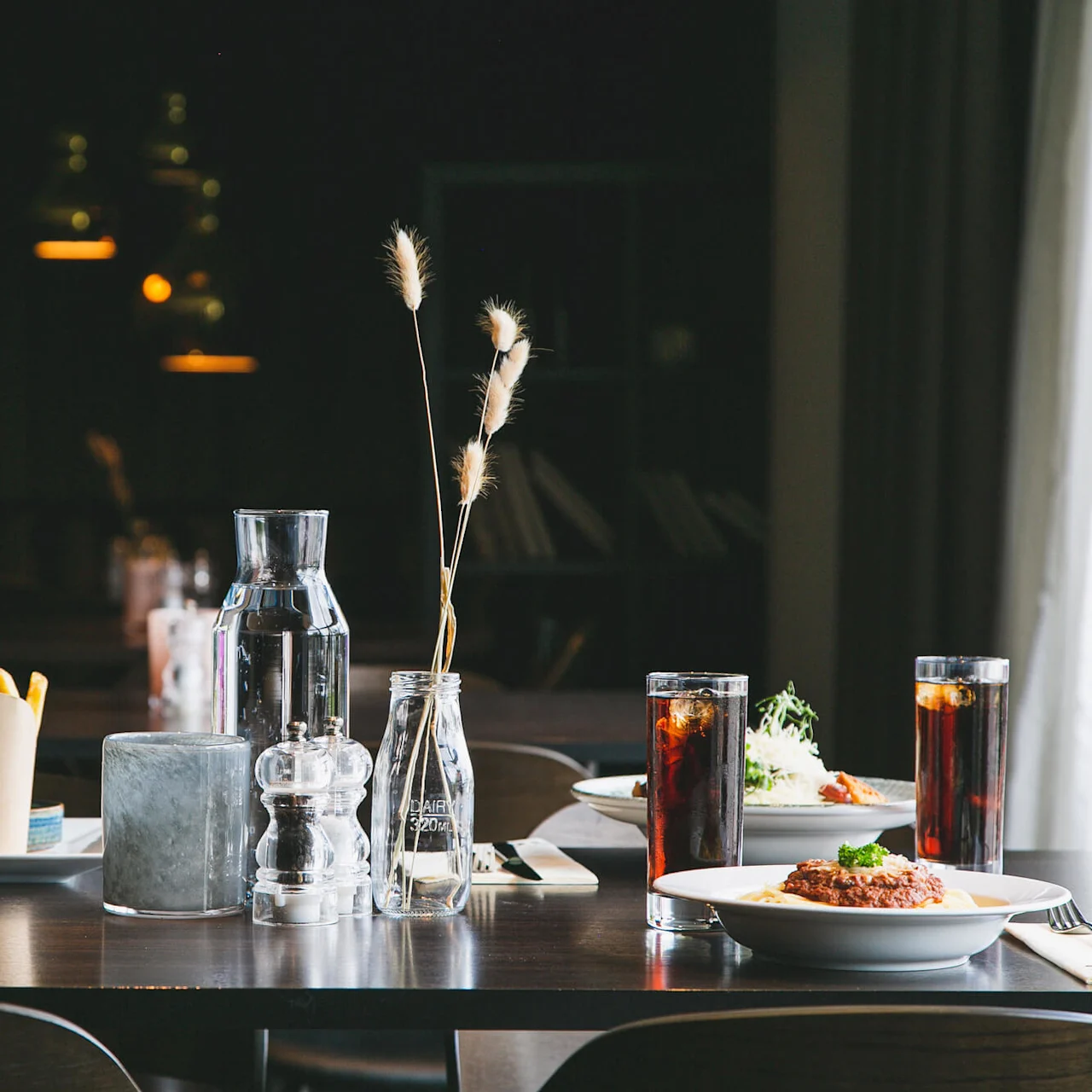 Et bord har en burger med pommes frites og drikkevarer, en pastaret og en vandkaraffel. Dæmpet belysning skaber en varm atmosfære i restaurantmiljøet.