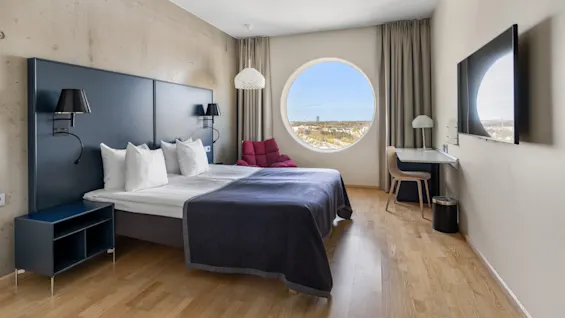 Et moderne hotelværelse med en stor seng, et rundt vindue med udsigt over byen, et skrivebord og et vægmonteret TV.