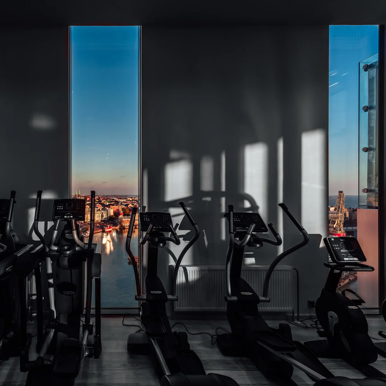Løbebånd og ellipsemaskiner står ubrugte i et fitnesscenter, med store vinduer, der giver en panoramaudsigt over byen ved solnedgang.