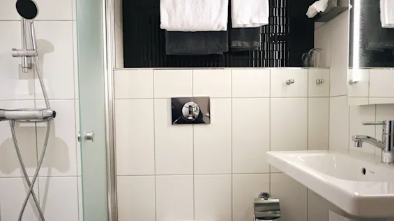 Toalett med en veggmontert flush knapp er plassert ved siden av en vask og dusj i et moderne bad, med hvite fliser og hengende håndklær.
