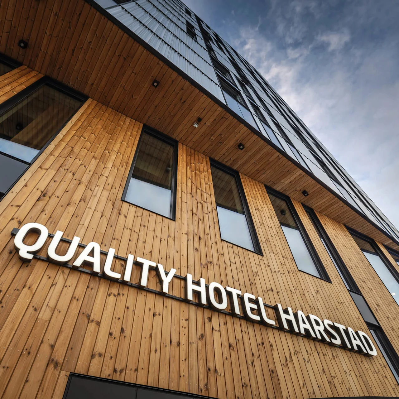 En træfacade med store vinduer viser skiltet "QUALITY HOTEL HARSTAD" mod en skyet himmel.
