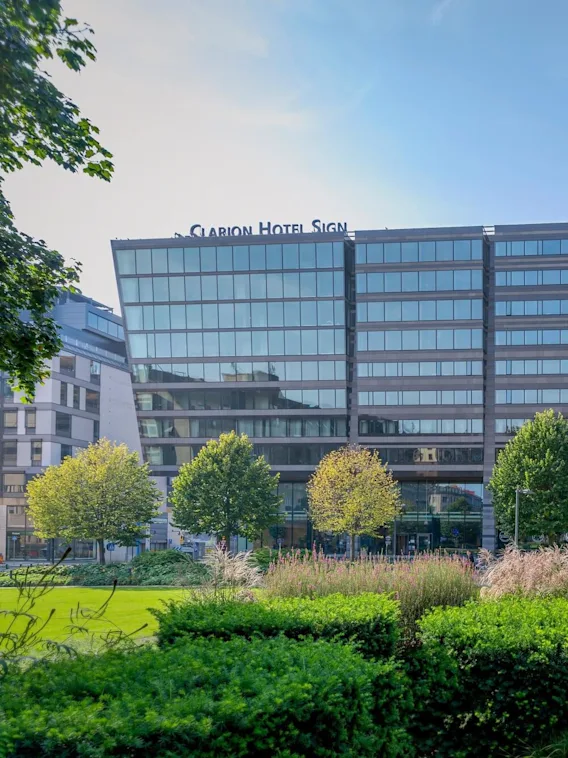 A modern, glass-fronted hotel building labeled "Clarion Hotel Sign" stands behind neatly arranged trees and bushes in an urban park setting.