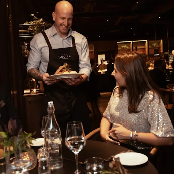 En servitør serverer en tallerken mat til en smilende gjest ved et restaurantbord