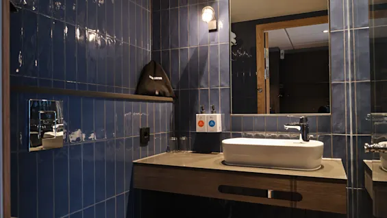 Et moderne badeværelse på Quality Hotel Match med blå fliser, en hvid håndvask, spejl og en hårtørrerpose på en hylde.