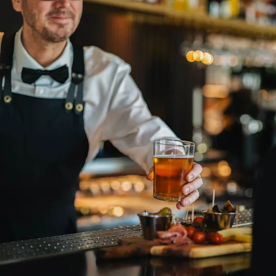Bartender in a bow tie serves a glass of beer to a person at a bar. A wooden board with snacks like tomatoes and olives is on the counter.