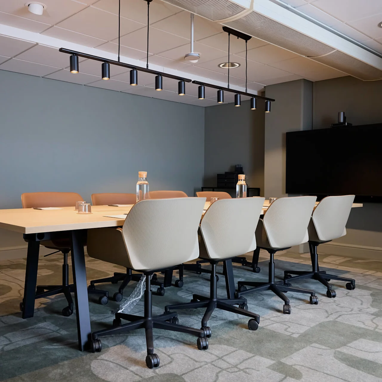 A conference room with a long table surrounded by beige chairs. Above, a black linear light fixture hangs. A large screen is mounted on the wall.