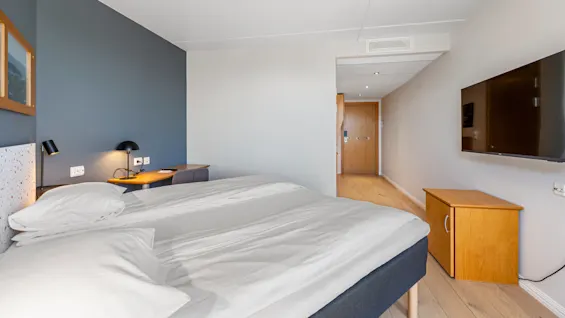 A tidy hotel room featuring a double bed, a work desk with a lamp, and a wall-mounted TV, in a contemporary style.