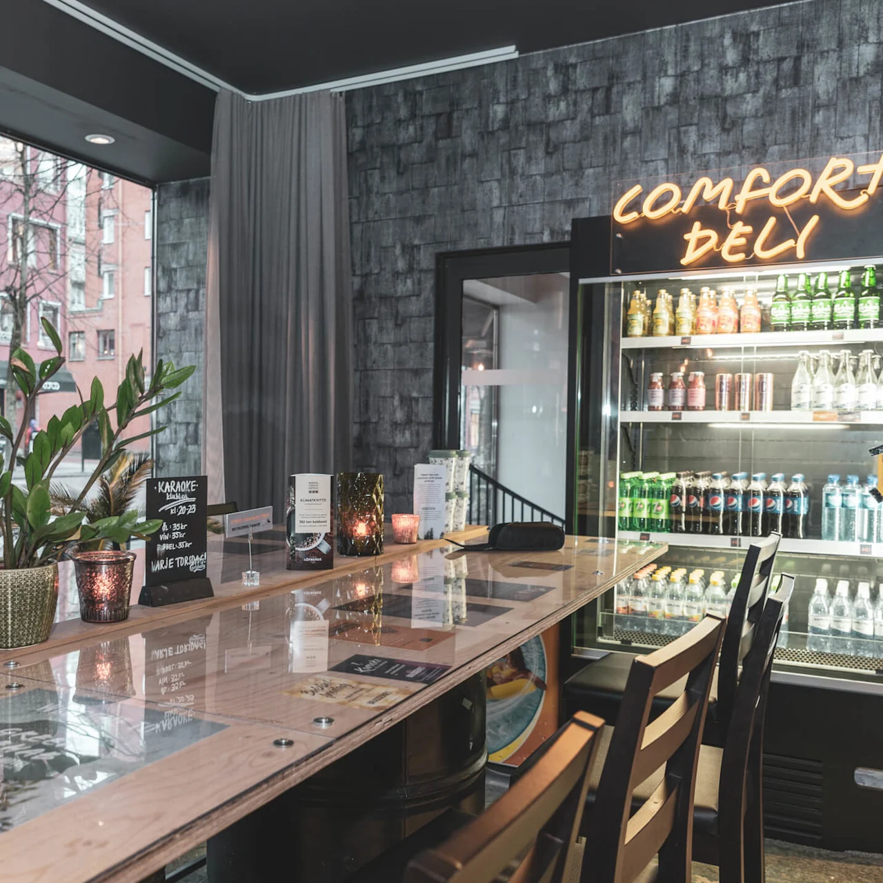 Et moderne deli-interiør med et fælles træbord, dekorative planter og et køleskab med drikkevarer. Neonsskilt læser "COMFORT DELI."