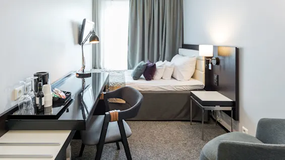 En seng prydelig oppredet med puter, plassert mot et vindu med lukkede gardiner, omgitt av et skrivebord med lampe og drikkevarer i et ryddig hotellrom.
