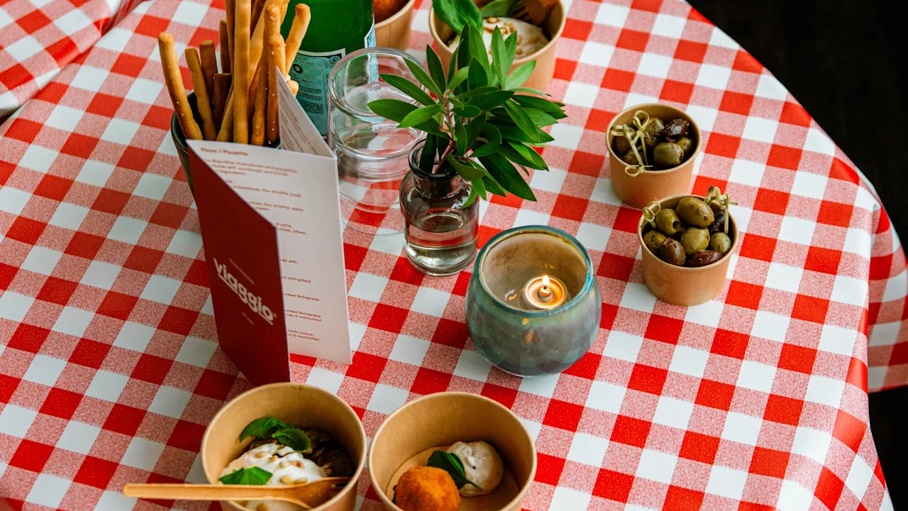 Rödvit-rutigt bordskläde med brödpinnar, flaskvatten, oliver, växt, ljus och två skålar med mat; omgiven av meny och glas på trägolv. Text på meny: "klassig".