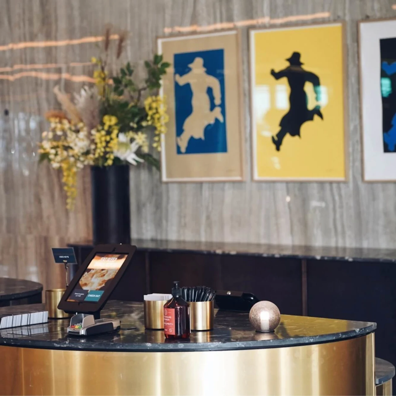 En resepsjonsdisk viser to digitale nettbrett ved siden av pyntegjenstander, plassert i en moderne, elegant lobby med tre silhuettmalerier på veggen og en vase med gule blomster.