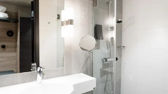 Tvättställ står under en spegel i ett modernt badrum med dusch med glasdörrar, grå kakel och en stark vägglampa.