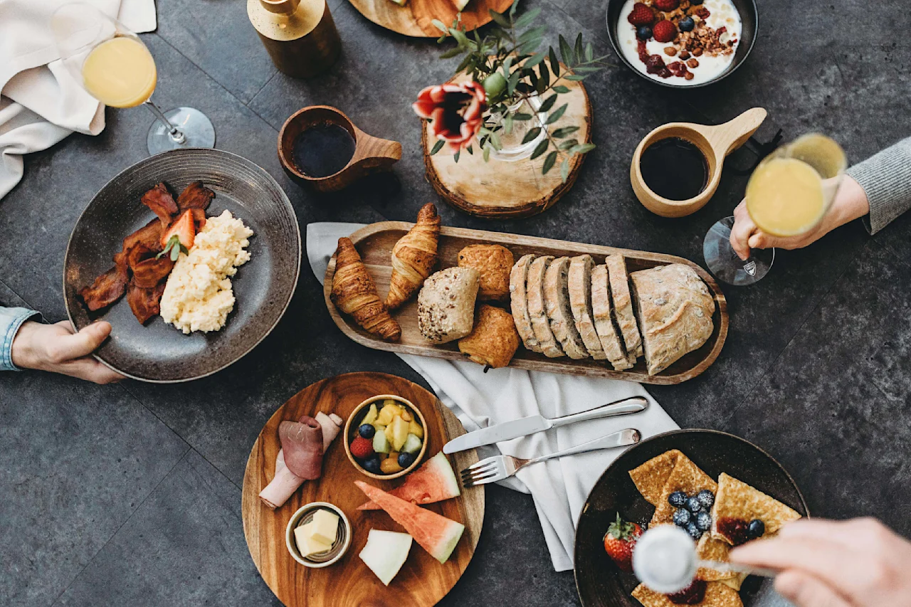 Tallerkener med morgenmadsmad, herunder bacon, æg, croissanter og frugter, er arrangeret på et mørkt bord med kaffe- og juiceglas, der skaber en brunchstemning. Hænder holder tallerkener og glas.