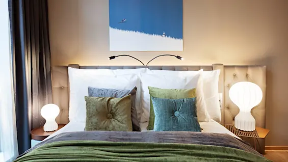 Bed adorned with plush green and gray pillows rests against a beige headboard; two modern lamps shine softly on side tables, framed by sheer curtains and abstract blue artwork above.