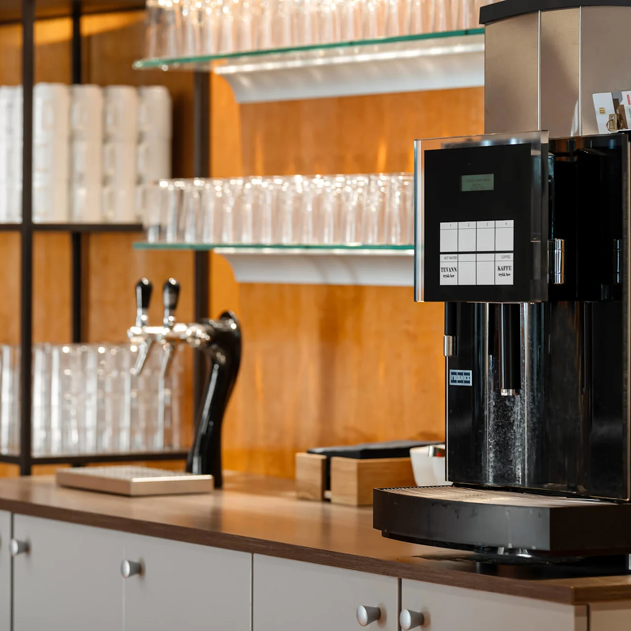Automatisk kaffemaskine står på en bordplade, omgivet af stablede glas og hylder med hvide kopper. Tekst på maskinens display: "Tee/Kaffee."