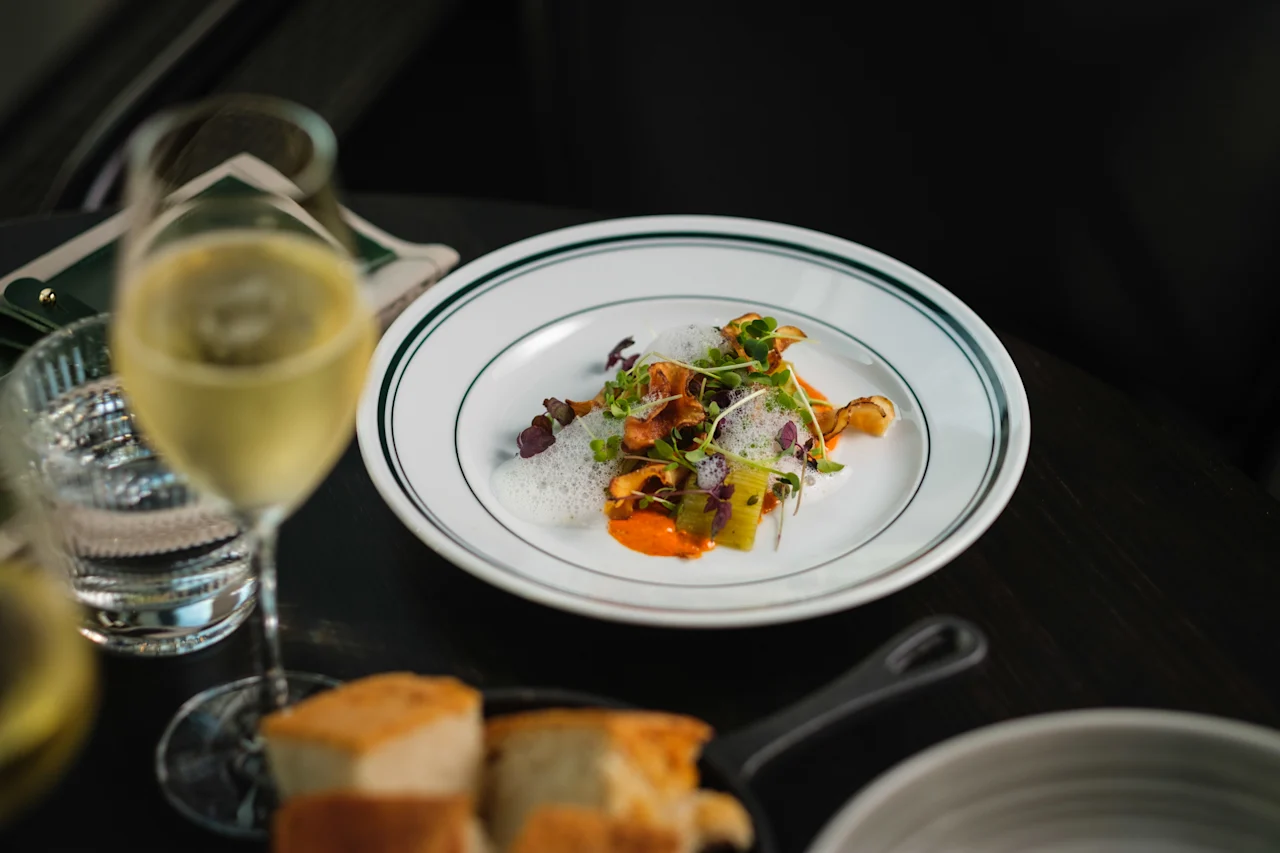 En gourmeträtt med färgglada grönsaker och skum ligger elegant på en vit tallrik, åtföljd av ett glas vitt vin och bröd, placerad på ett mörkt bord.