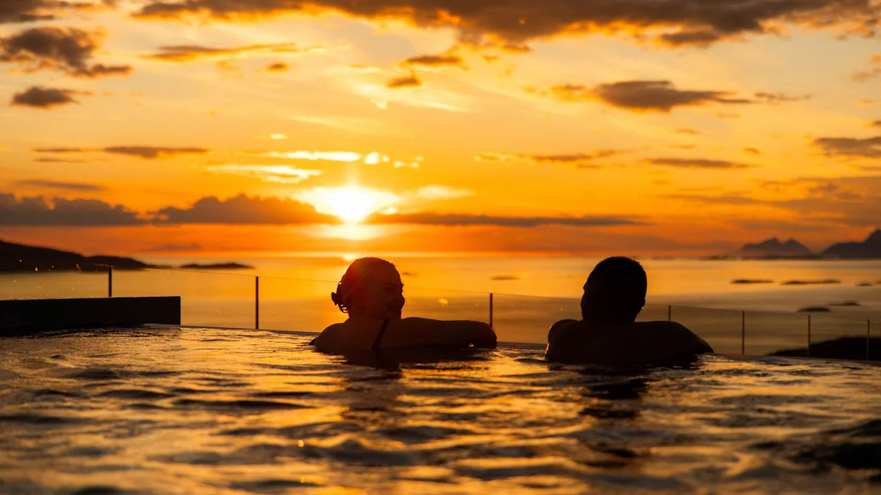 To personer slapper af i en pool og ser en farverig solnedgang over et stort hav. Silhuettskyer og fjerne landskaber giver dybde til den fredelige scene.