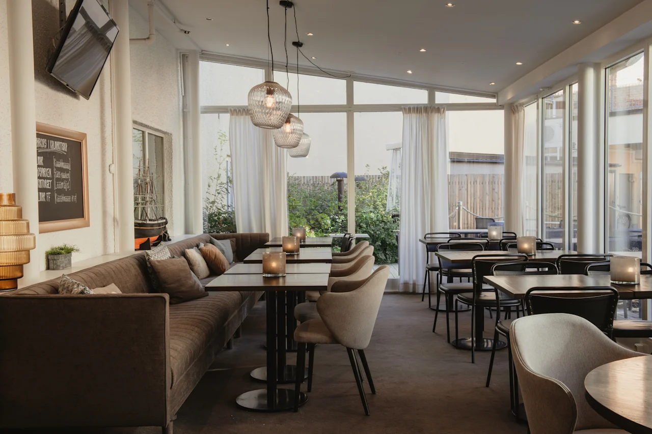 En restaurant med en lang sofa, borde og store vinduer. En menutavle viser BURGERS DRINKS SNACKS DESSERT.