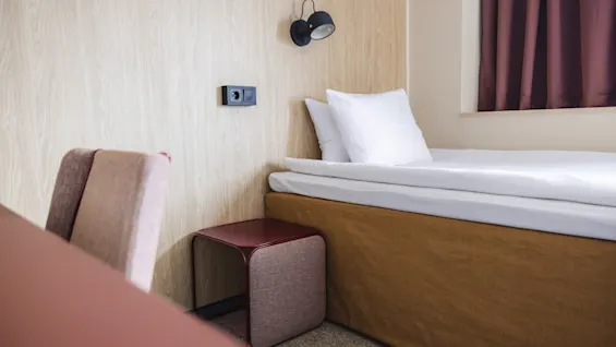 Bed with white pillows; resting quietly; in a minimalist room with wood-panel wall, purple chair, side table, wall light, and curtained window.