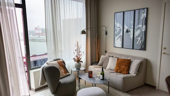 Bekväm sittgrupp med soffa och fåtöljer, dekorerade med kuddar och en växt, vid ett fönster med utsikt över vatten och byggnader, vilket skapar en mysig, modern miljö.