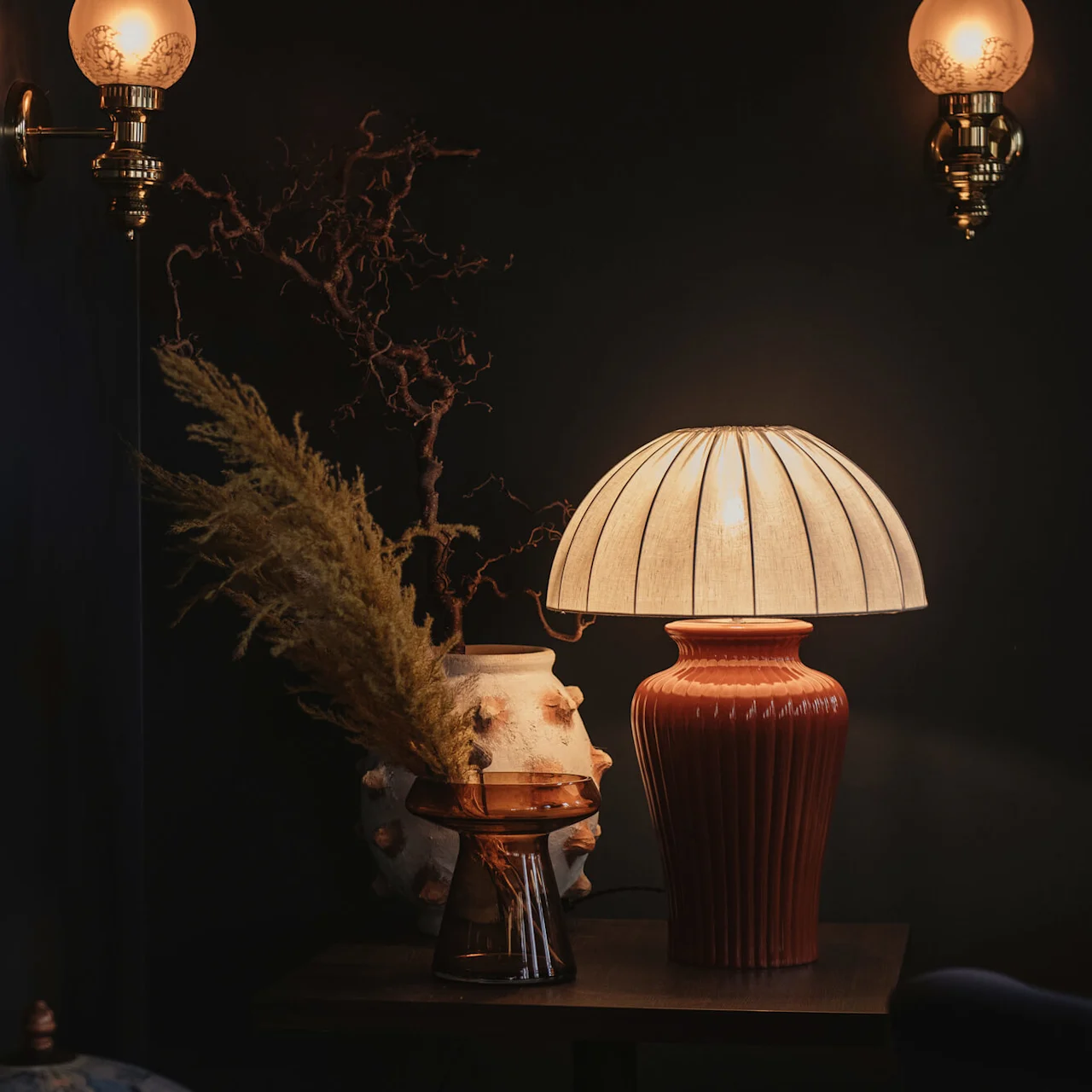 A table lamp with a pleated shade softly illuminates a dark room, surrounded by decorative vases and dry plants. Elegant wall sconces provide additional lighting above a framed painting.