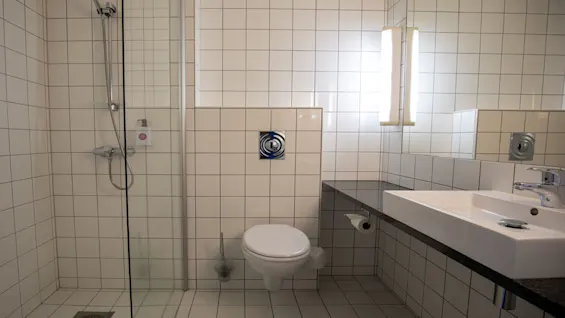 Et badeværelse med vægmonteret toilet og brusebad indkapslet af glas. En håndvask med spejl til højre, mod hvide flisebelagte vægge og gulv, godt oplyst miljø.