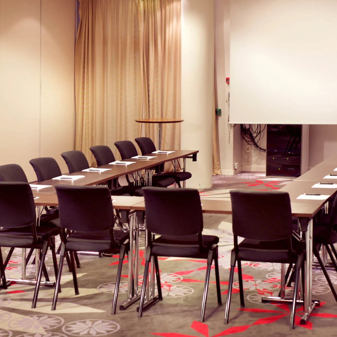 Conference room arranged with chairs and tables, papers on tables, with a projector screen and flip chart, and patterned carpet on the floor.