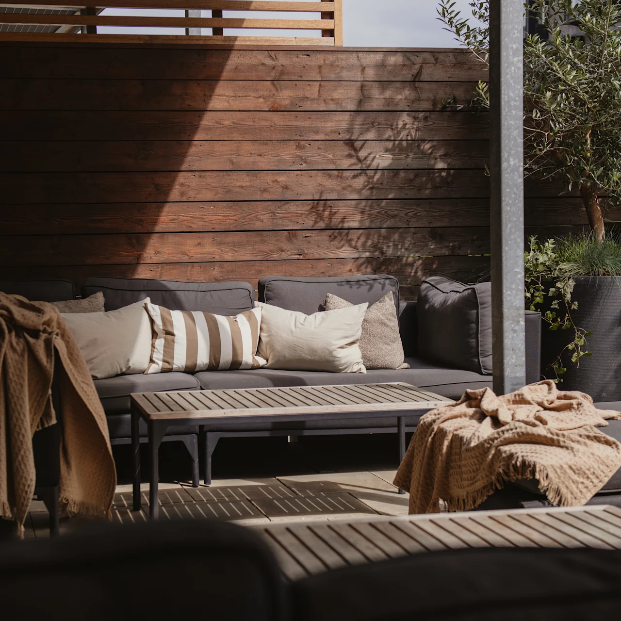 A cozy outdoor seating area featuring gray couches with beige cushions, wooden tables, and decorative blankets, set against a wooden fence and large potted plants. 