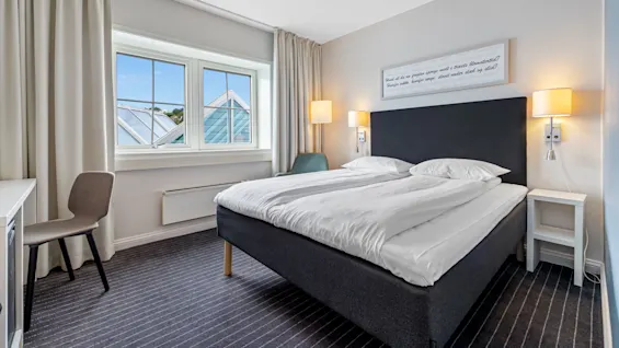 A tidy hotel room with an unmade bed, facing a window with geometric roof details; a framed text is above the bed.