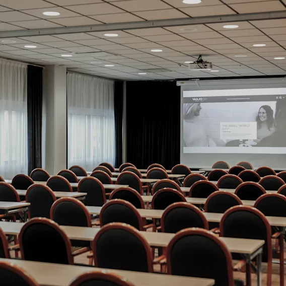 Klassrum med rader av bord och stolar vända mot en projiceringsskärm som visar en presentation. Rummet är väl upplyst med stora fönster och en takmonterad projektor.