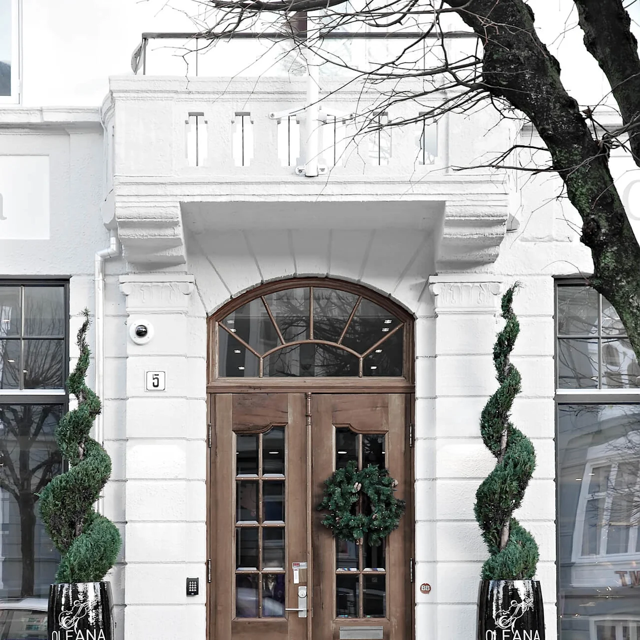 A wooden double door, adorned with a Christmas wreath, stands between two spiral topiary plants in black pots labeled "OLEANA". The white building has large windows and a balcony.