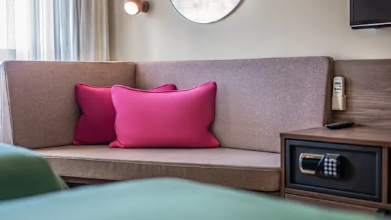 A brown corner sofa with two pink cushions is placed in a cozy room. A wooden side table with a safe, remote, and wall light adds functionality.