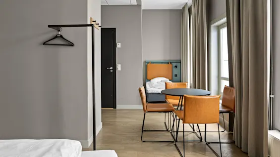 Siisti hotellihuone, jossa on pyöreä pöytä ja neljä nahkatuolia ikkunan vieressä. Vuode valkoisilla liinavaatteilla ja seinälle asennetut ripustimet lisäävät yksinkertaisuutta ja eleganssia.