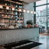 En moderne bar har en flisebeklædt disk med hængende lys, fyldte hylder og planter; ved siden af store vinduer med udsigt over en bygade. Tekst på en måtte lyder: "SIT BACK, RELAX AND ENJOY."