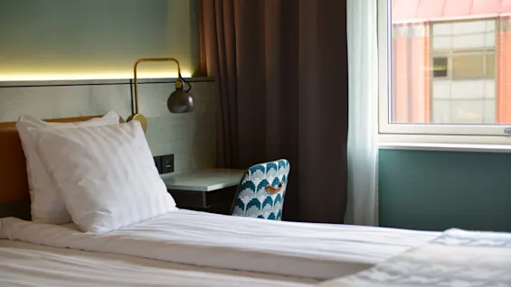 En pænt redt seng med hvidt sengetøj står ved siden af et vindue. En mønstret stol vender mod et lille skrivebord med lampe. Gardinerne dækker delvist vinduet.