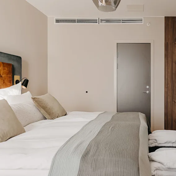 A neatly made bed with beige pillows and a grey throw rests against a wall, facing a mounted TV. A wooden wardrobe and a door are visible beside it.