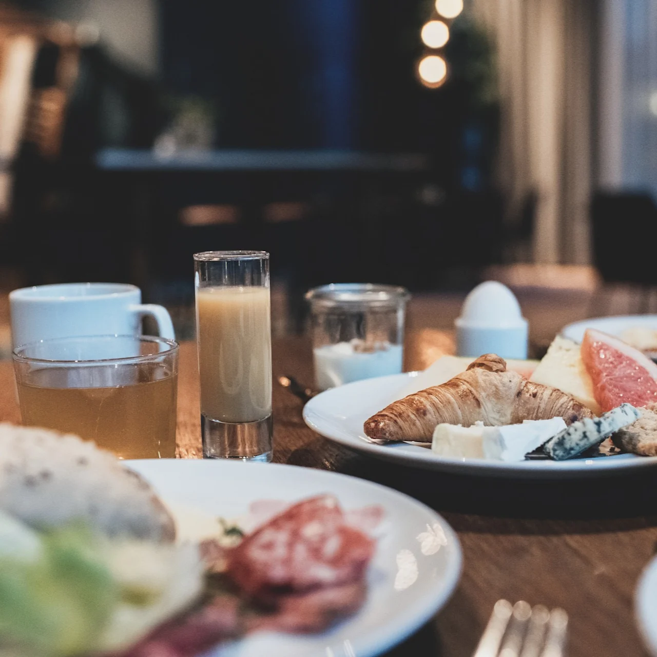 Ett frukostbord visar tallrikar med en croissant, ost, grapefrukt och kallskuret. Tillhörande drycker är apelsinjuice och te. Bakgrunden har en mjukt upplyst matsal.