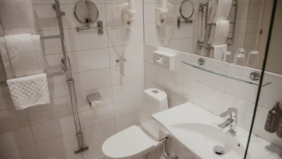 Toilet i et flisebelagt badeværelse ved siden af en vask med vandhane. To hårtørrer er monteret på væggen, og håndklæder hænger på en håndklædetørrer; glas står på en glashylde.