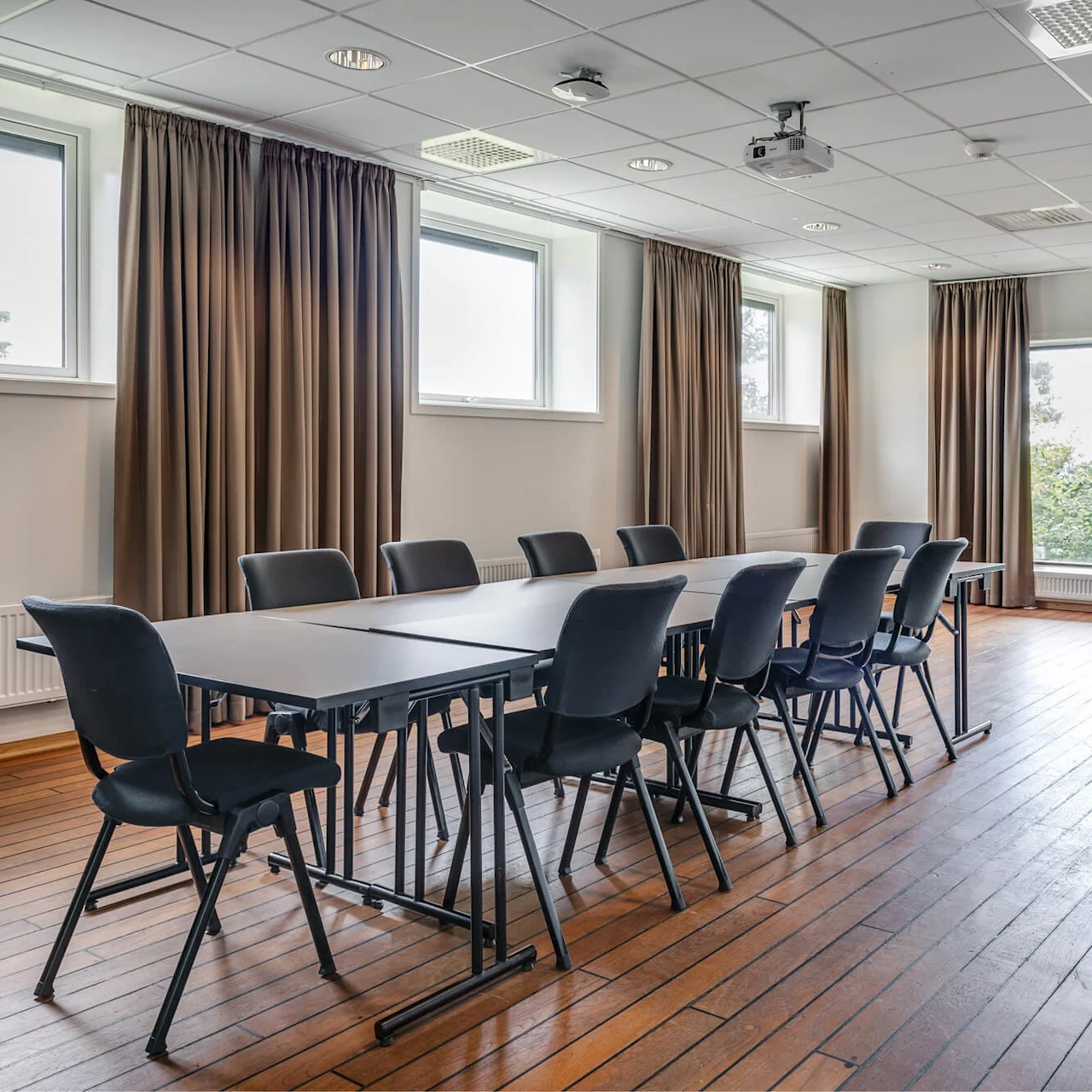 Et langt bord med svarte stoler er satt opp i et konferanserom. Rommet har store vinduer med brune gardiner, tregulv og en takmontert projektor.
