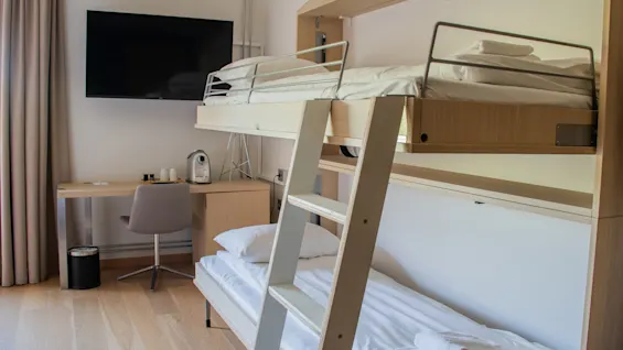 Bunk bed with a ladder in a tidy room; lower bed has white sheets. Nearby, a desk holds a coffee machine, chair, and beneath a wall-mounted TV. Curtains frame the scene.